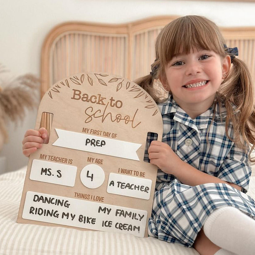 Four-year-old girl sitting on bed and holding Hello Fern Back to School non-custom board in her school uniform.