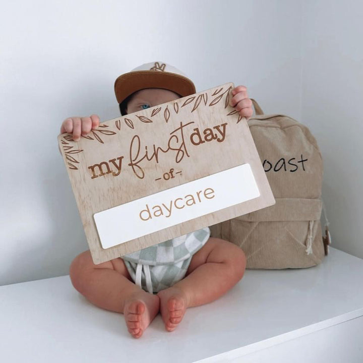 Toddler with backpack holding Hello Fern "my first day of" school board with the word "daycare" added to the white acrylic panel.