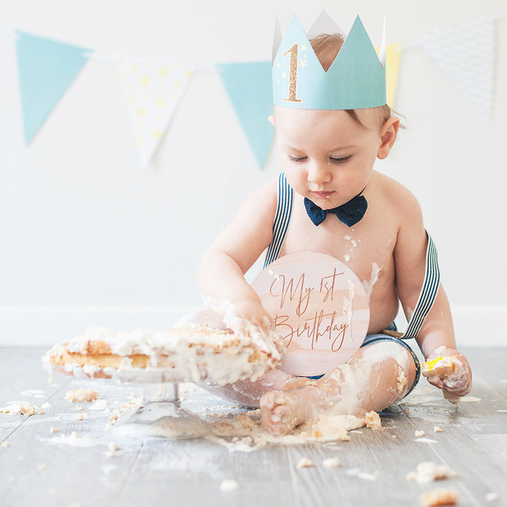 Baby boy messily playing with birthday cake with Hello Fern "My 1st Birthday" wooden plaque on lap.