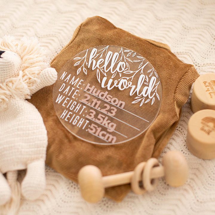 Hello Fern clear acrylic 'Hello Word' plaque with white wording and alphabet stickers lying on baby blanket with baby items surrounding.