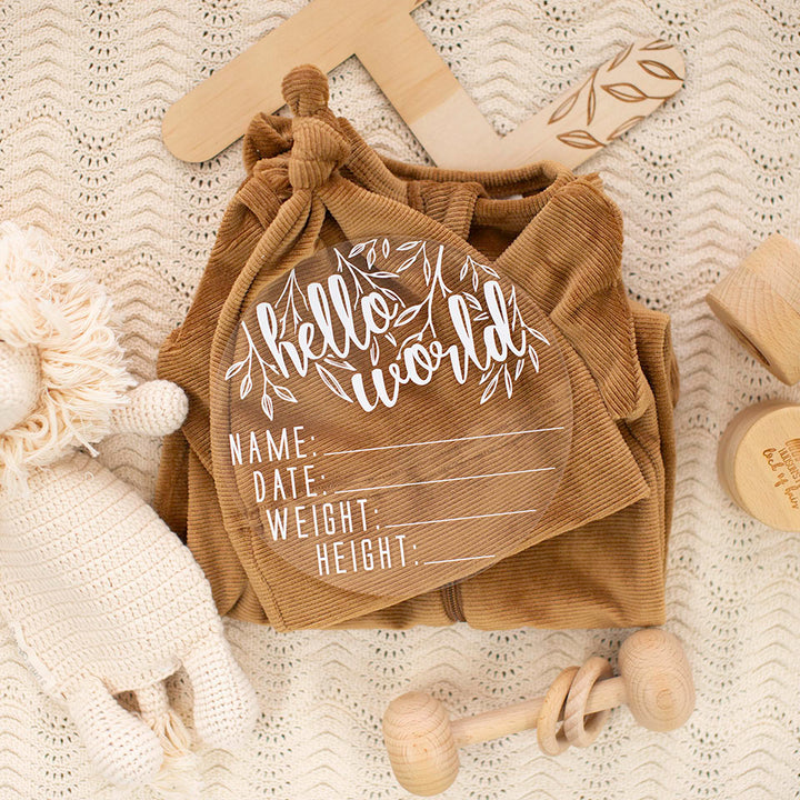 Top view of Hello Fern clear acrylic 'Hello Word' birth announcement plaque with white wording and spaces for baby's name, birth date, weight, and height lying on blanket with baby items surrounding.