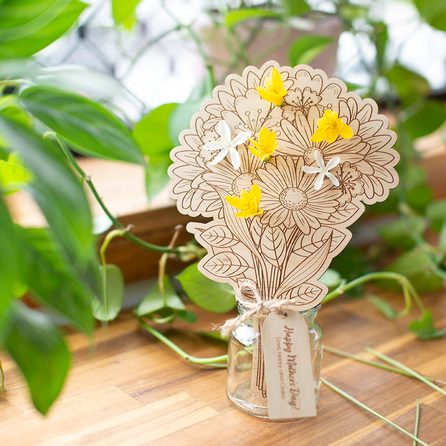 Hello Fern wooden flower bouqet in glass jar sitting next to sunlit window ledge with plants and wildflowers.