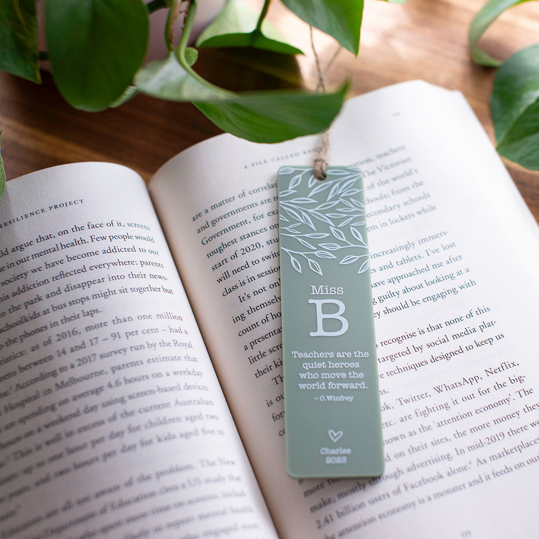 Photo of open book with custom printed Hello Fern sage acrylic teacher bookmark in portrait orientation, natural light, and plants in the background.