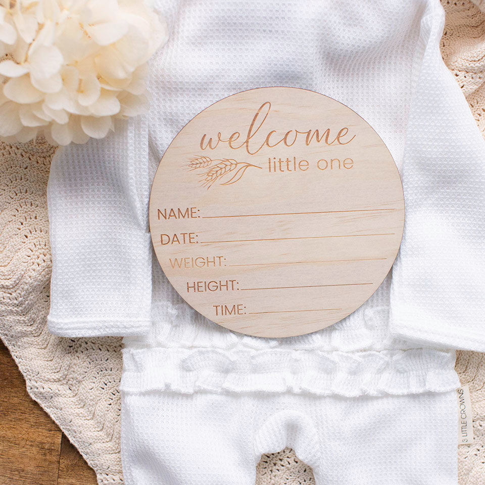 Overhead view of 'welcome little one' country-themed baby birth announcement disc including spaces for name, birth date, weight, height, and time. on baby outfit with blanket and dried flowers.