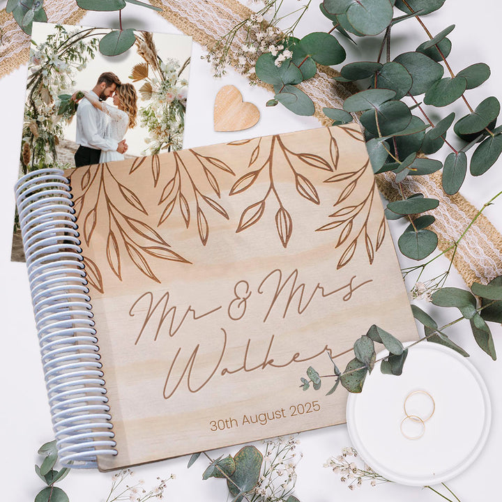 Hello Fern wooden wedding photo album with custom wording etched on front cover. Shown as flat lay with wedding photo of bride and groom, wedding rings, and plants.