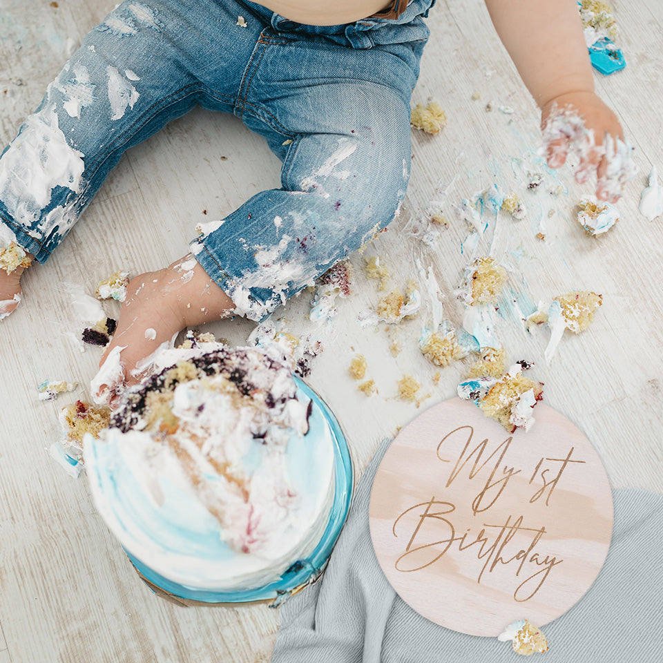 Top view of little boy's legs and hands with messy birthday cake smeared all over floor and Hello Fern "My 1st Birthday" announcement plaque.
