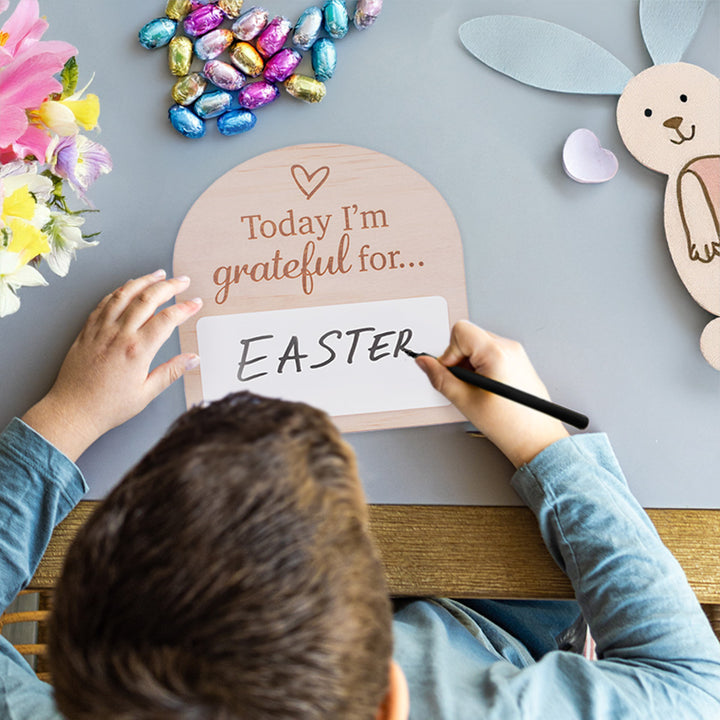 Little boy writing on "Today I am grateful for..." Hello Fern mini affirmation message plaque on table top with chocolate Easter eggs, bunny, and flowers.
