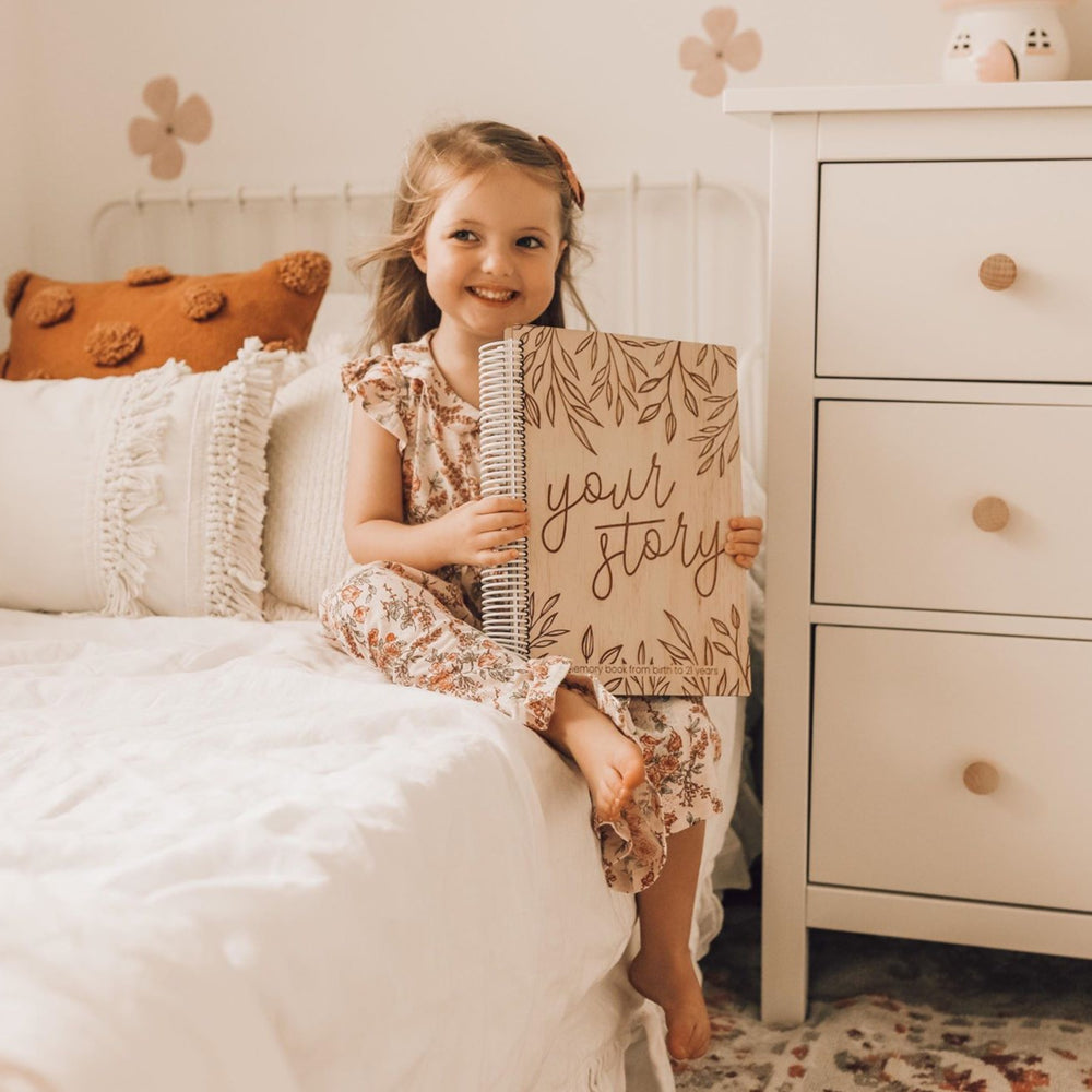 Little girl sitting on bed and smiling, whilst holding Hello Fern's "your story" non-custom wooden baby memory book.