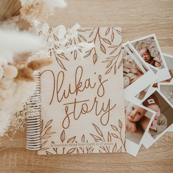 Flatlay showing 'Iluka's story' etched into cover of custom Hello Fern wooden baby memory book with polaroid baby photos and dried flowers.