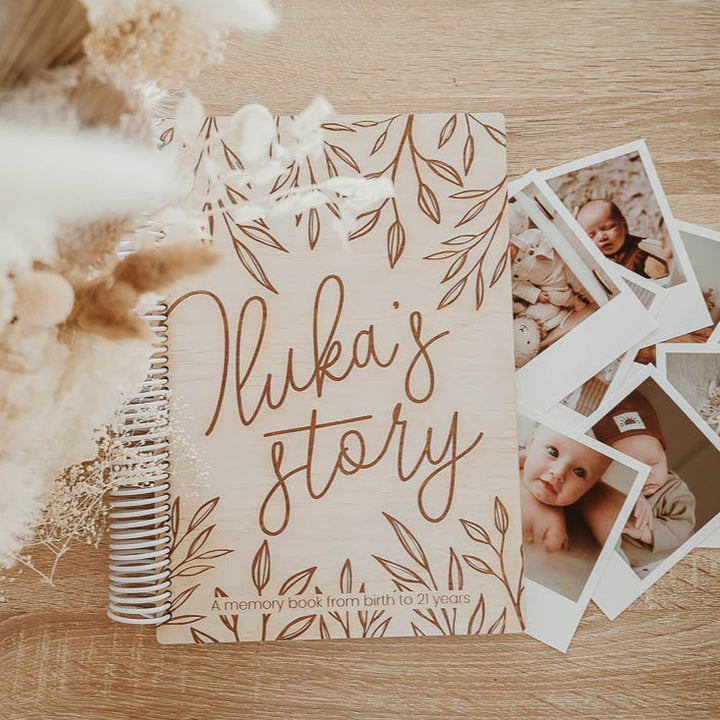 Flatlay showing 'Iluka's story' etched into cover of custom Hello Fern wooden baby memory book with polaroid baby photos and dried flowers.