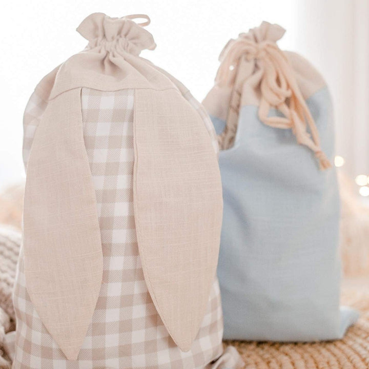 Two cotton Easter sacks (beige gingham, and blue) with bunny ears and custom wooden letter name plaque sitting on table.