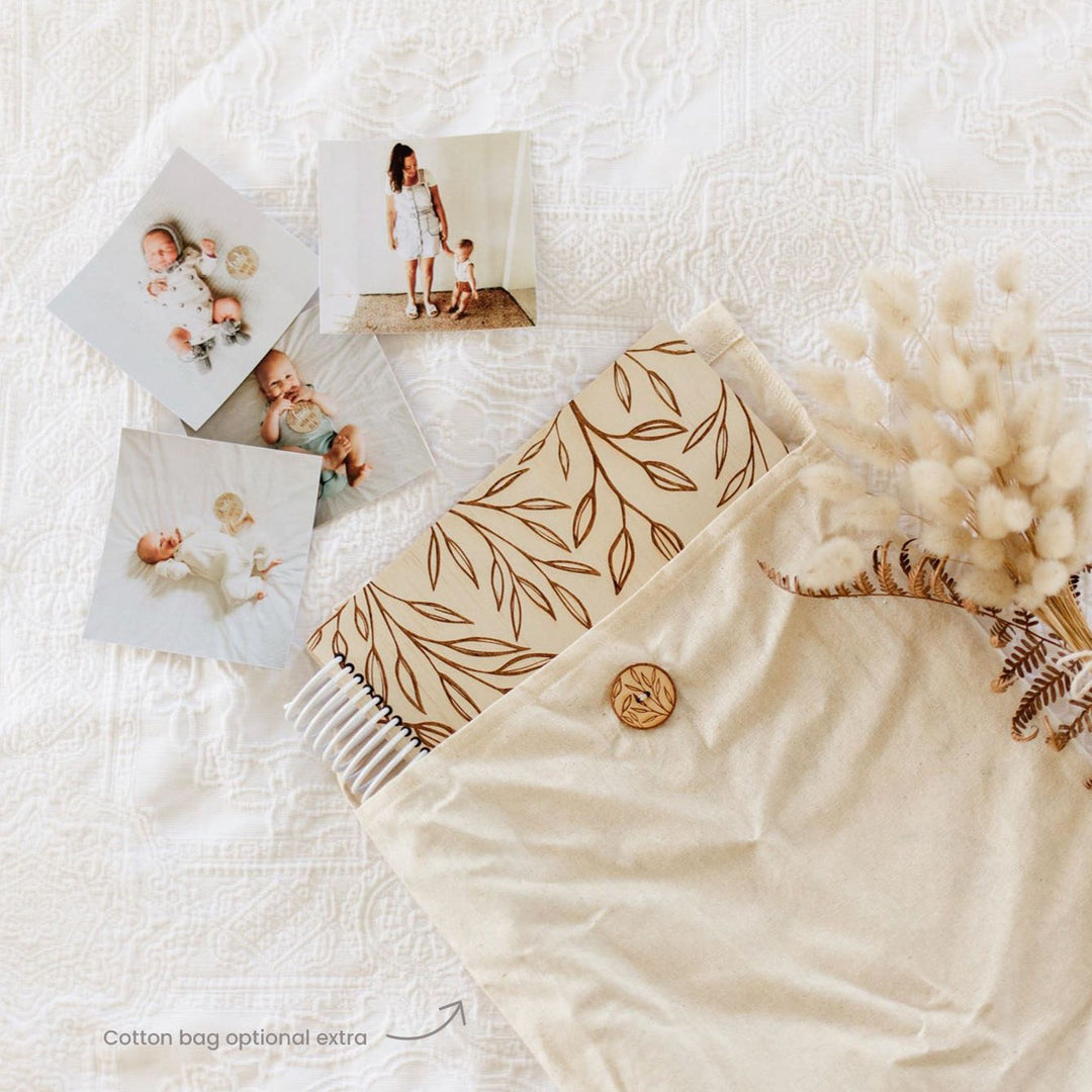 Hello Fern wooden baby memory book shown inside handmade cotton bag with baby photos scattered around.