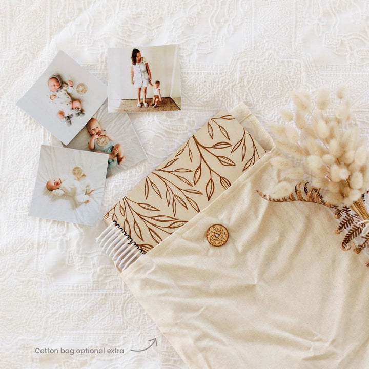 Hello Fern wooden baby memory book shown inside handmade cotton bag with baby photos scattered around.
