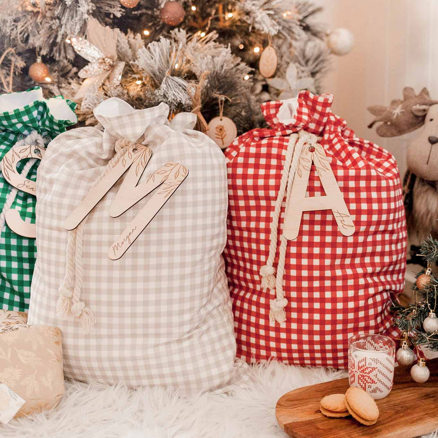 Set of three Christmas Santa sacs in front of a Christmas tree. Green sac with letter 'S' wooden plaque, brown sac with letter 'M' wooden plaque, and red sac with letter 'A' wooden plaque.