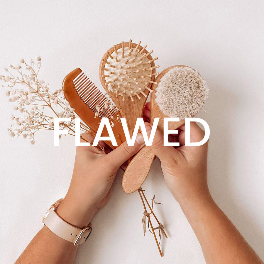 Hands holding a Hello Fern non-custom brush set including two wooden baby brushes and a comb with word FLAWED
