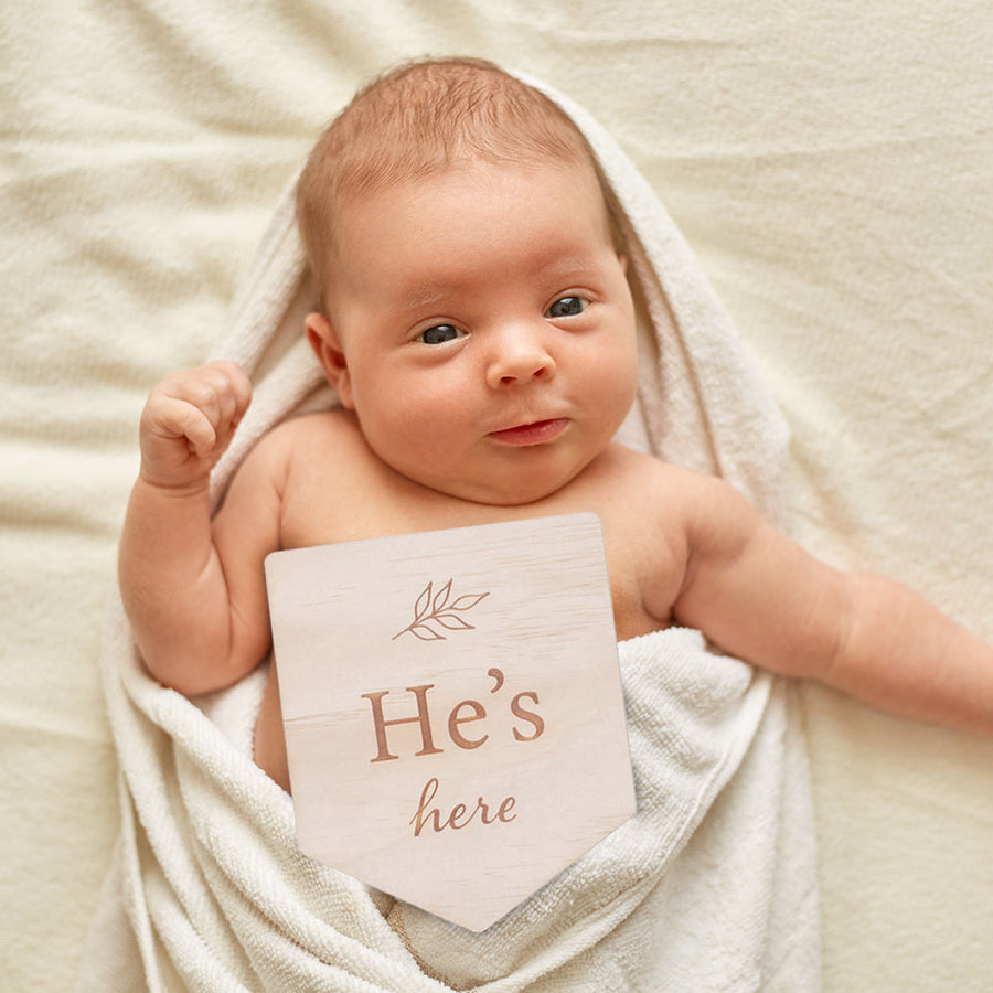 Newborn baby lying in towel after bath with Hello Fern double-sided wooden birth announcement plaque etched with the words "He's here" and a simple leaf graphic.