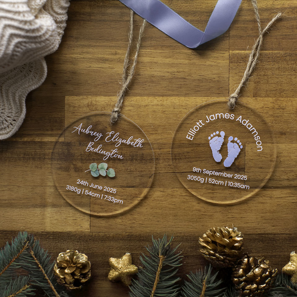 Two Hello Fern clear acrylic Christmas ornament baubles with printed baby names birth details on a wooden surface with decorative elements.