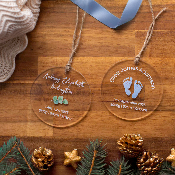 Two Hello Fern clear acrylic Christmas ornament baubles with printed baby names birth details on a wooden surface with decorative elements.