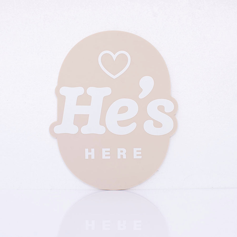 Hello Fern beige acrylic birth announcement plaque in oval shape with printed words "He's Here" and small heart graphic.