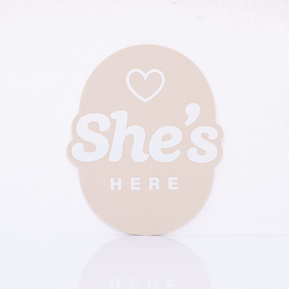 Hello Fern beige acrylic birth announcement plaque in oval shape with printed words "She's Here" and small heart graphic.