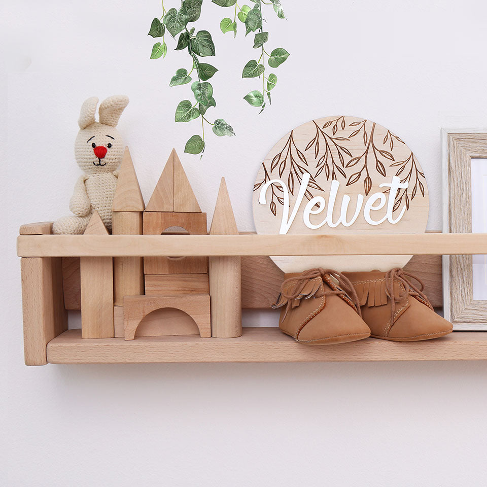 Children's room shelf decor with Hello Fern wooden and acrylic baby name plaque displayed next to wooden blocks, stuffed toy, and baby mocassins.