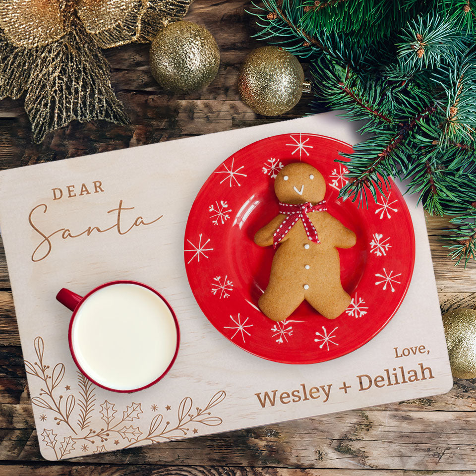 Overhead view of gingerbread man cookie on red plate and milk in mug for Santa placed on Hello Fern custom wooden Santa snack board with pine and Christmas decorations.