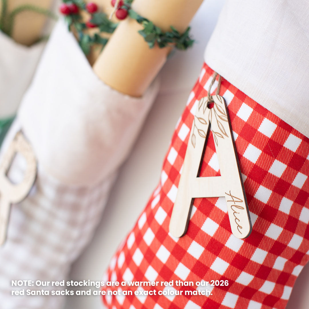 A close-up of a red and white gingham Christmas stocking with a wooden monogram letter 'A' hanging from it, alongside other holiday stockings in the background.