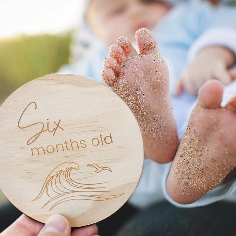 Baby's feet with sand, holding a Hello Fern wooden disc marking 'Six months old' with beach design.