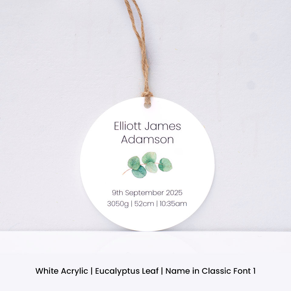 Round white acrylic Hello Fern Christmas ornament with eucalyptus leaves and personalised with baby name and birth details on a light grey background.