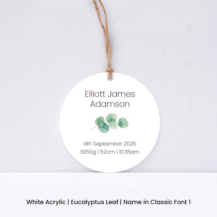 Round white acrylic Hello Fern Christmas ornament with eucalyptus leaves and personalised with baby name and birth details on a light grey background.
