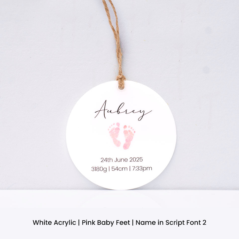 Round white acrylic Hello Fern Christmas ornament with pink baby feet print and personalised with baby name and birth details on a light grey background.
