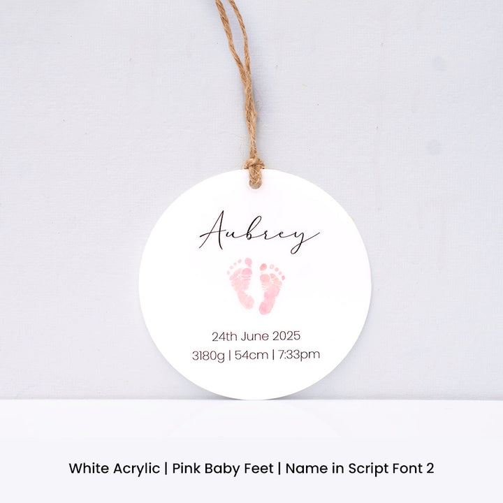 Round white acrylic Hello Fern Christmas ornament with pink baby feet print and personalised with baby name and birth details on a light grey background.