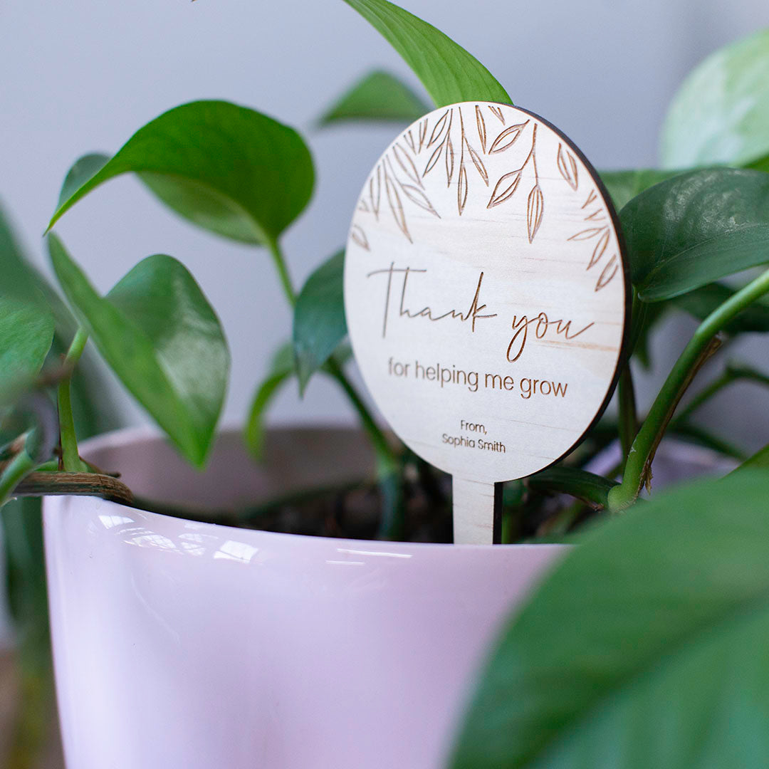Teacher gift wooden circle-shaped plant stake with 'Thank you for helping me grow' message and custom sign-off shown in potted plant.