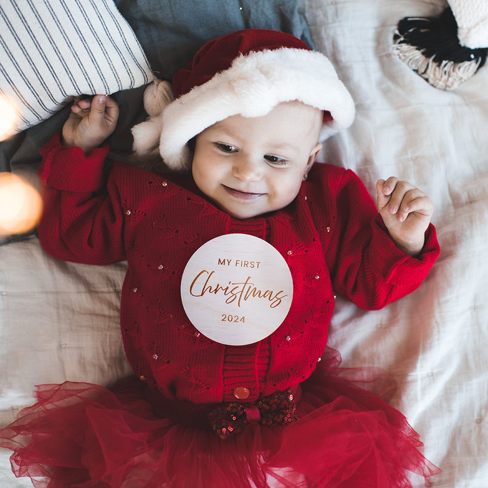 Cute baby girl with Santa hat and holiday dress lying on bed with Hello Fern non-custom wooden "My first Christmas" keepsake disc.