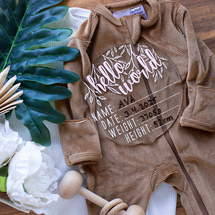 Top view of Hello Fern clear acrylic 'Hello Word' birth announcement plaque with pink wording and filled-out spaces for baby's name, birth date, weight, and height lying on baby girl clothes.