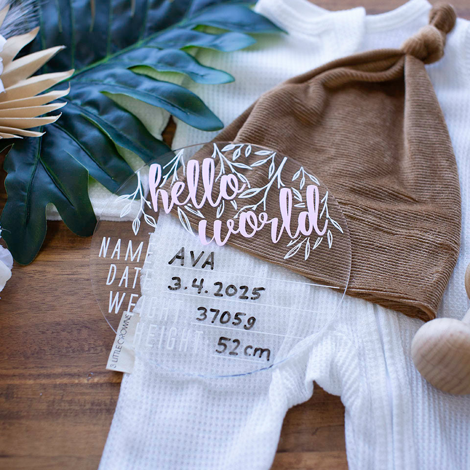 Hello Fern clear acrylic 'Hello Word' birth announcement plaque with pink wording and filled-out spaces for baby's name, birth date, weight, and height lying on baby clothes.
