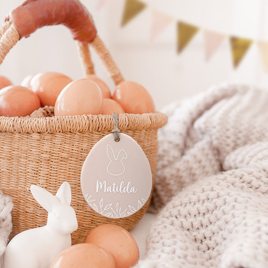 Hello Fern beige acrylic custom Easter basket tag hanging on basket full of eggs with Easter decor in background.