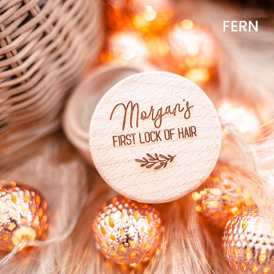 Closeup of Hello Fern baby's first lock of hair wooden trinket box engraved with custom name showing lid open and surrounded by ornamental lights.