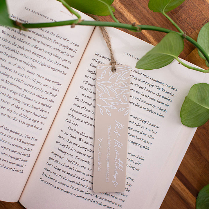 Top view open novel with custom printed Hello Fern beige acrylic teacher bookmark in landscape orientation and plants in the background.