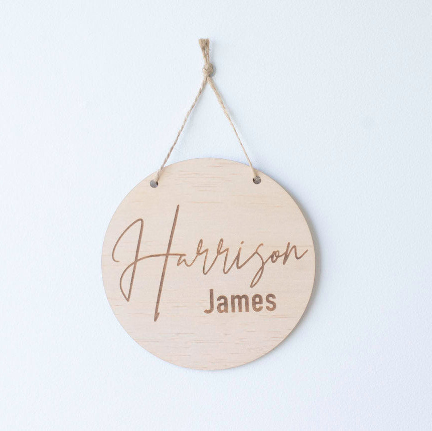 Round wooden plaque by Hello Fern with 'Harrison James' engraved, hanging with twine on a white wall.