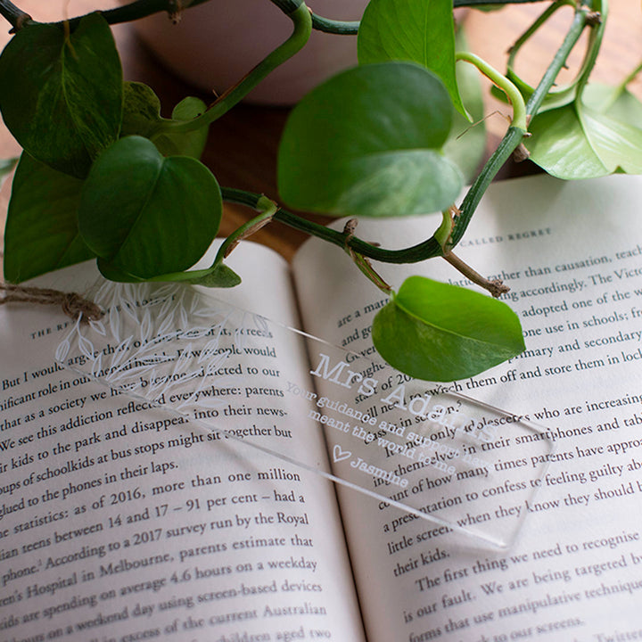 Photo of open book on wooden table with custom printed Hello Fern clear acrylic teacher bookmark in landscape orientation and plants in the background.