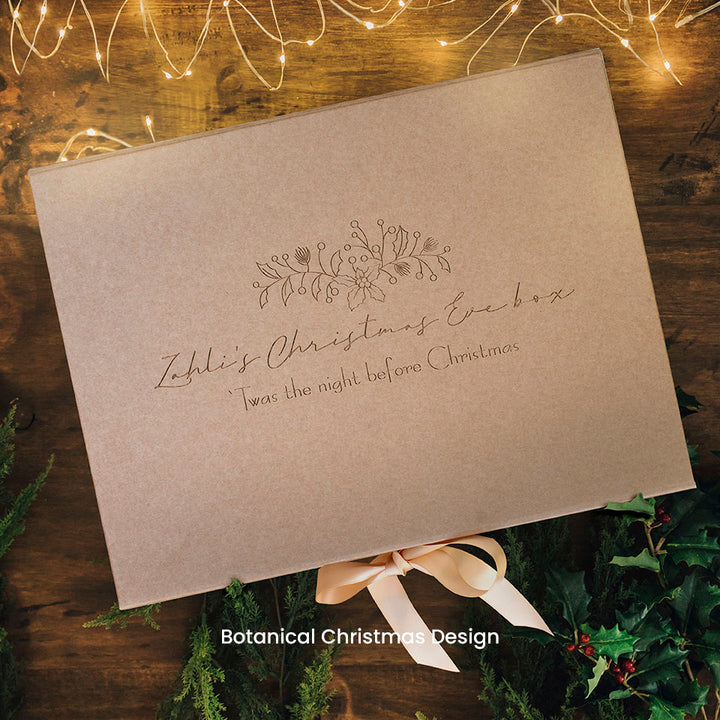 Hello Fern kraft paper Christmas Eve keepsake gift box with botanical design, decorative text, and bow on a wooden surface with Christmas lights and decorations.