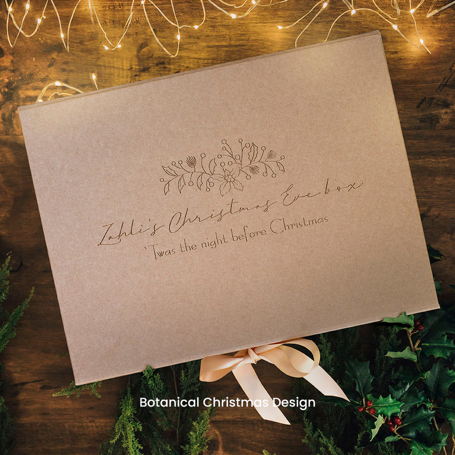 Hello Fern kraft paper Christmas Eve keepsake gift box with botanical design, decorative text, and bow on a wooden surface with Christmas lights and decorations.