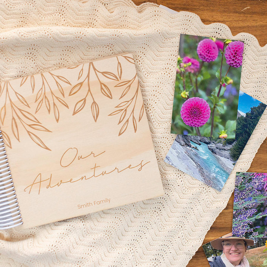 Hello Fern custom wooden photo album with personalised engraved front cover and beautiful flower and family holiday photos scattered on table.