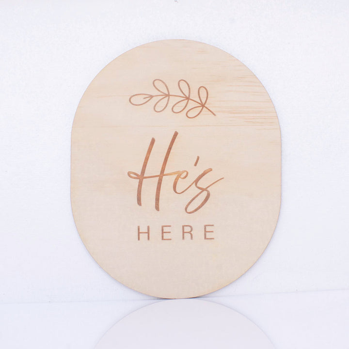 Hello Fern double-sided wooden birth announcement plaque in an oval shape displaying the side with the words "He's here" with a small fern graphic.