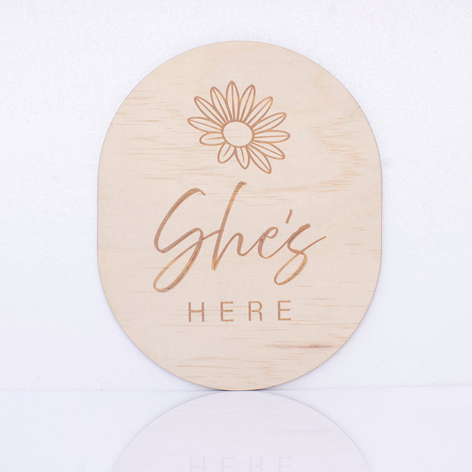 Hello Fern double-sided wooden birth announcement plaque in an oval shape displaying the side with the words "She's here" with a simple flower graphic.