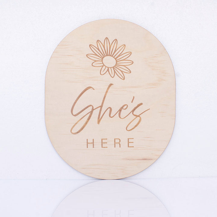 Hello Fern double-sided wooden birth announcement plaque in an oval shape displaying the side with the words "She's here" with a simple flower graphic.