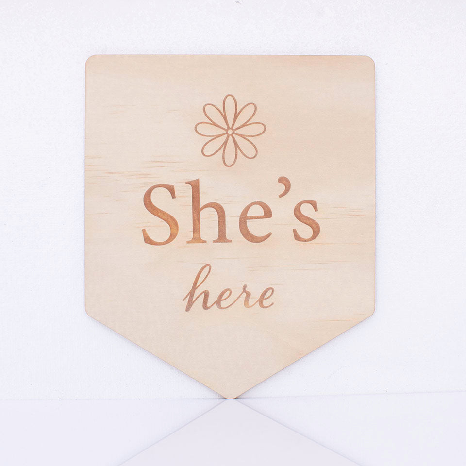 Hello Fern double-sided wooden birth announcement plaque in a pennant shape displaying the side with the words "She's here" with a simple flower graphic.