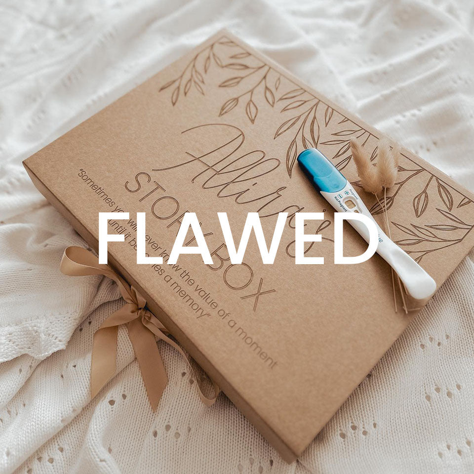 A beige cardboard keepsake box with a custom etched name on the lid, alongside a pregnancy test and decorative elements. The box is designed to hold a wooden baby book and is accompanied by a ribbon for closure with 'FLAWED' text overlay