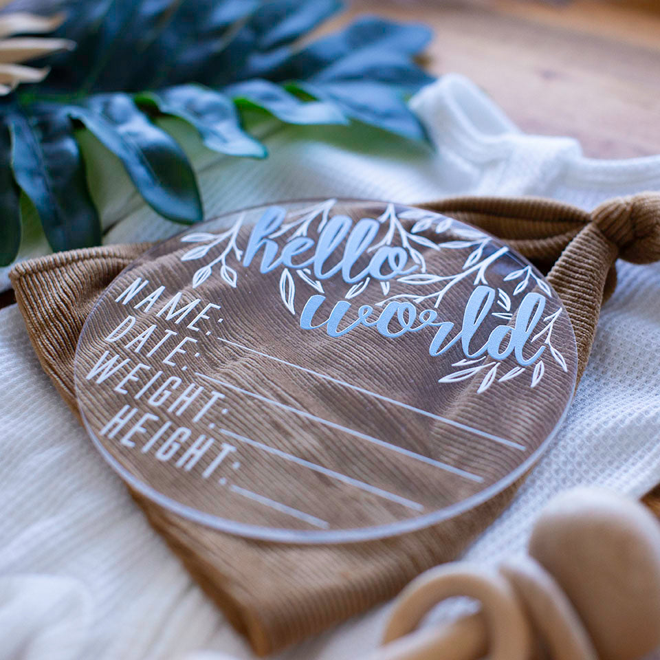 Closeup view of Hello Fern clear acrylic 'Hello Word' birth announcement plaque with blue wording and blank spaces for baby's name, birth date, weight, and height lying on baby items.